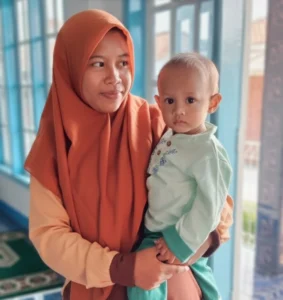 Seorang ibu tersenyum mendampingi anaknya di rumah sederhana, mencerminkan peran ibu sehat dan bahagia dalam membangun keluarga tangguh.